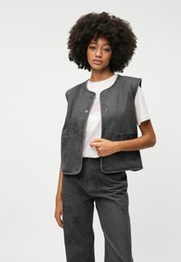 Grå denimvest med rund halslinning, trykknapper, to lommer og subtile tonale trykk. Brukt over en hvit t-skjorte med grafikk.