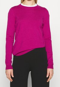 Fuchsia Strickpullover mit langen Ärmeln, rundem Halsausschnitt und gerippten Bündchen. Getragen über einem weißen Hemd, kombiniert mit schwarzen Hosen. Weiche Textur.