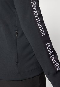 Donkergrijze sportjack met een gladde textuur, aansluitend ontwerp en een zwart paneel met de tekst "Peak Performance" langs de mouw.