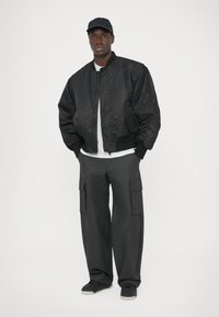 Juoda bombonešio stiliaus striukė su užtrauktuku priekyje, struktūruotais rankogaliais ir šoninėmis kišenėmis; derinama su laisvomis tamsiomis „cargo“ kelnėmis su daugybe kišenių. Juoda kepurė ir batai.