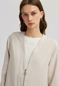 Lys beige zip-up blazer med dyb V-hals, lavet af et tekstureret, letvægtsstof. Har minimalistisk design og sølvhardware.