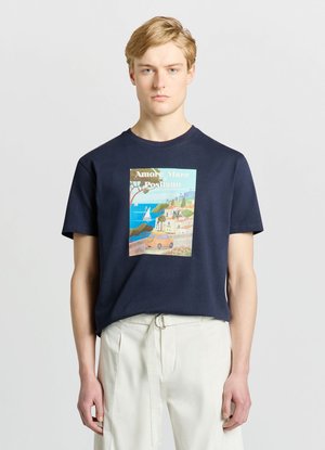 Jeune homme aux cheveux clairs porte un t-shirt bleu marine avec un imprimé coloré d'une ville balnéaire et un pantalon blanc avec ceinture en tissu sur fond uni.