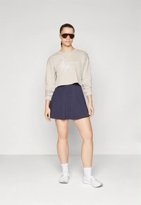 Bėgianti smėlio spalvos megztinio palaidinė su grafiniu spaudiniu, derinama su tamsiai mėlyna skaterio tipo sijonu. Modelis dėvi oversized tipo akinius nuo saulės ir baltus sportinius batelius.