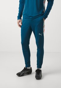 Teal sportbroeken van ademend materiaal, met een slim fit, zijzakken en een Puma-logo met een gemusterd ontwerp. Zwarte sportschoenen.