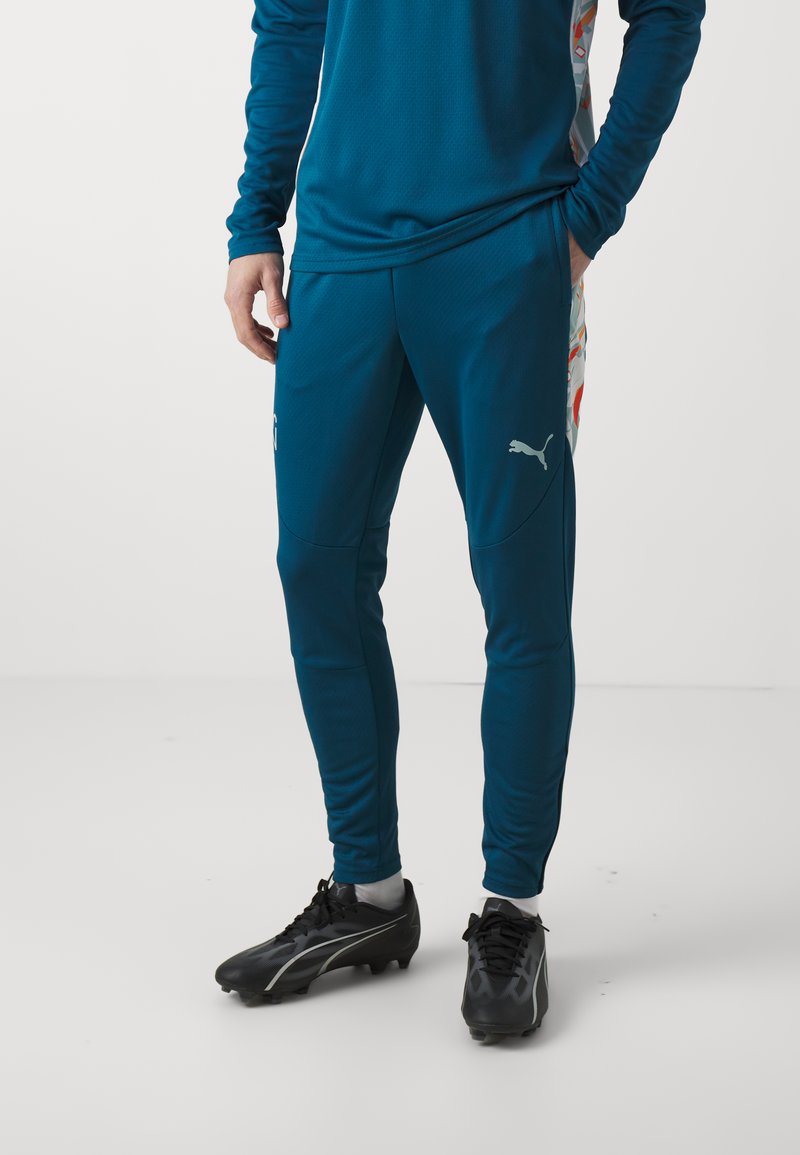Teal sportbroeken van ademend materiaal, met een slim fit, zijzakken en een Puma-logo met een gemusterd ontwerp. Zwarte sportschoenen.