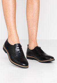Musta nahkakengät, joissa on sileä pinta, pyöreä kärki, koristeellinen brogue-yksitys sekä matala pinojalko. Varusteena nyöritettävä muotoilu.