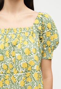 Femme portant une robe blanche avec imprimé citrons jaunes et feuilles vertes, avec un col carré et des manches courtes bouffantes avec détails smockés.