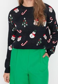 Czarny dziergany sweter z świątecznym wzorem przedstawiającym bałwanki, choinki i laski cukrowe w kolorach czerwonym, zielonym i białym. Noszony z zielonymi spodniami.
