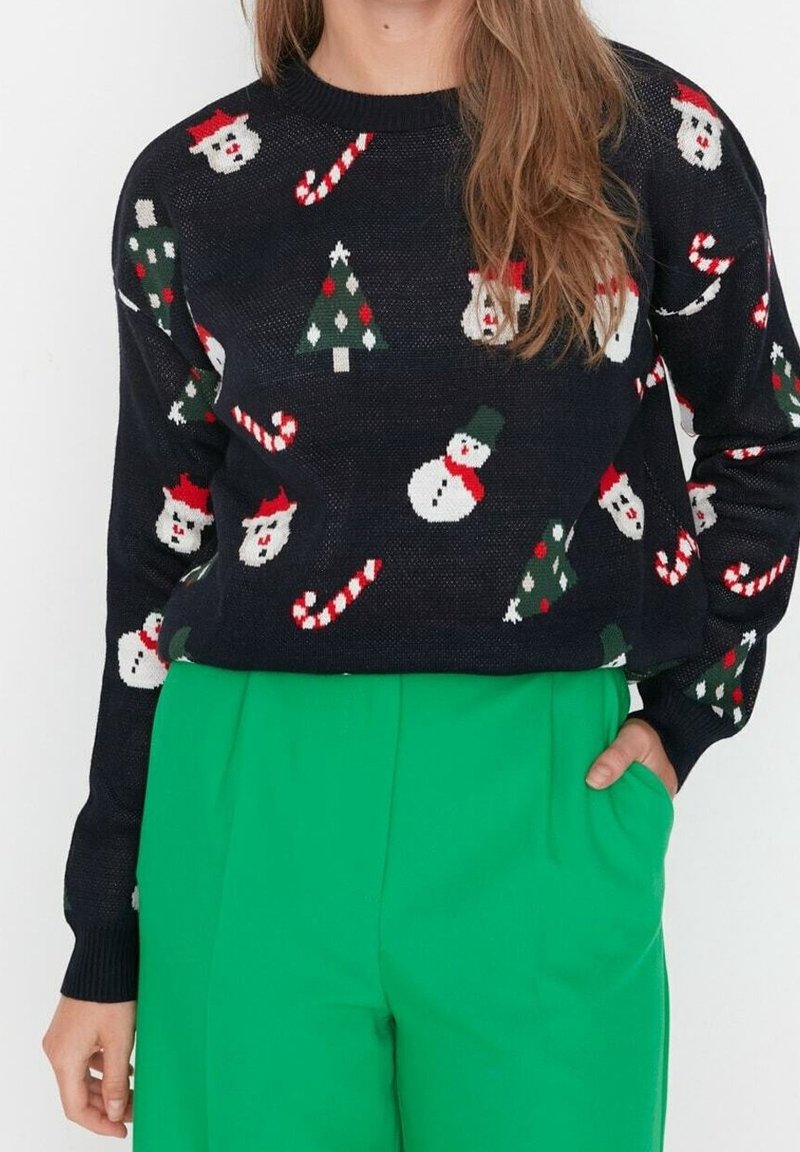 Czarny dziergany sweter z świątecznym wzorem przedstawiającym bałwanki, choinki i laski cukrowe w kolorach czerwonym, zielonym i białym. Noszony z zielonymi spodniami.