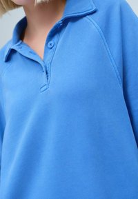 Blauwe polo met een kraag, een placket met twee knopen en een textuurstof met een gladde afwerking. Zichtbare stikseldetails bij de hals en schouders.