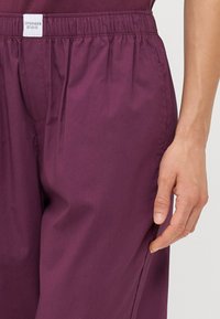 Pantalons bordeaux à taille élastique avec poches latérales, texture lisse et détail froncé au niveau de la taille. Étiquette visible.