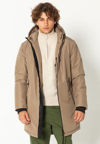 Beige wasserdichter Parka mit Kapuze, schwarzen Reißverschlüssen und einer Fronttasche. Glatte Textur, mittellanges Design, über einem leichten Pullover getragen.
