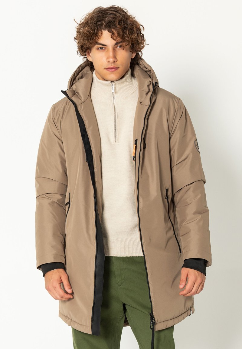 Beige wasserdichter Parka mit Kapuze, schwarzen Reißverschlüssen und einer Fronttasche. Glatte Textur, mittellanges Design, über einem leichten Pullover getragen.