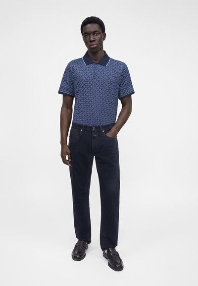 Homme debout, main dans la poche, portant un polo bleu à motifs, un jean foncé et des chaussures en cuir noires, sur un fond blanc uni.