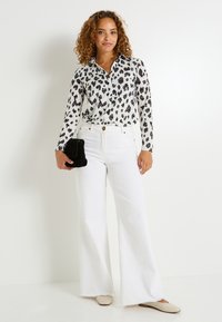 Blouse à manches longues avec des taches de léopard noires sur fond blanc, associée à un jean flare blanc taille haute et des ballerines de couleur claire.