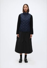 Ναυτικό γιλέκο με επένδυση και ψηλό γιακά, με φερμουάρ και υφή, φορεμένο πάνω από μια μαύρη μακριά φούστα και μαύρα παπούτσια.