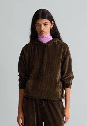 Donna con lunghi capelli scuri che indossa una felpa con cappuccio e pantaloni di velluto marrone, con un dolcevita viola chiaro sotto, in piedi davanti a uno sfondo semplice.