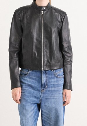 Personne portant une veste en cuir noire zippée avec col pressionné et un jean bleu taille haute sur un fond uni.