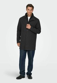 Ciemnoszary wełniany płaszcz z wysokim kołnierzem, zapinany z przodu na guziki, z bocznymi kieszeniami, zestawiony z niebieskimi dżinsami i czarnymi skórzanymi butami.