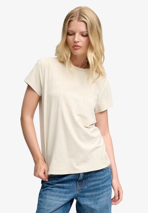 Mujer rubia con camiseta beige de manga corta y vaqueros azules, mirando hacia abajo con una mano sujetando el dobladillo de la camiseta.