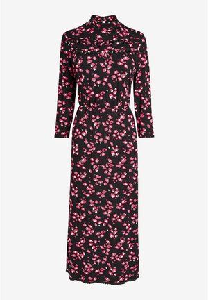 Femme portant une robe midi noire à imprimé floral rose avec des manches trois-quarts, associée à des baskets blanches, debout devant un fond vert.
