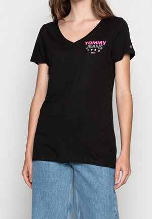 Kobieta w czarnej koszulce z dekoltem w serek z napisem "TOMMY JEANS 1985" w różowym i białym kolorze na piersi, w połączeniu z jasnoniebieskimi dżinsami.