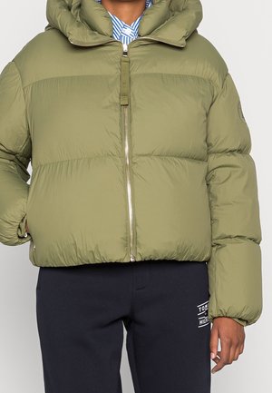 Person trägt eine olivgrüne Steppjacke mit durchgehendem Reißverschluss, kombiniert mit dunkelblauen Tommy Hilfiger Jogginghosen.
