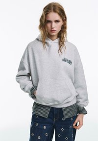 Felpa grigia con cappuccio, tasca frontale e testo ricamato. Abbinata a una camicia a quadri bianca e nera sotto e jeans in denim con dettagli di occhielli metallici.