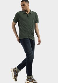 Polo shirt vert foncé, jeans bleus et chaussures de sport multicolores. Le polo a un col et un petit logo, tandis que les jeans ont une coupe classique.