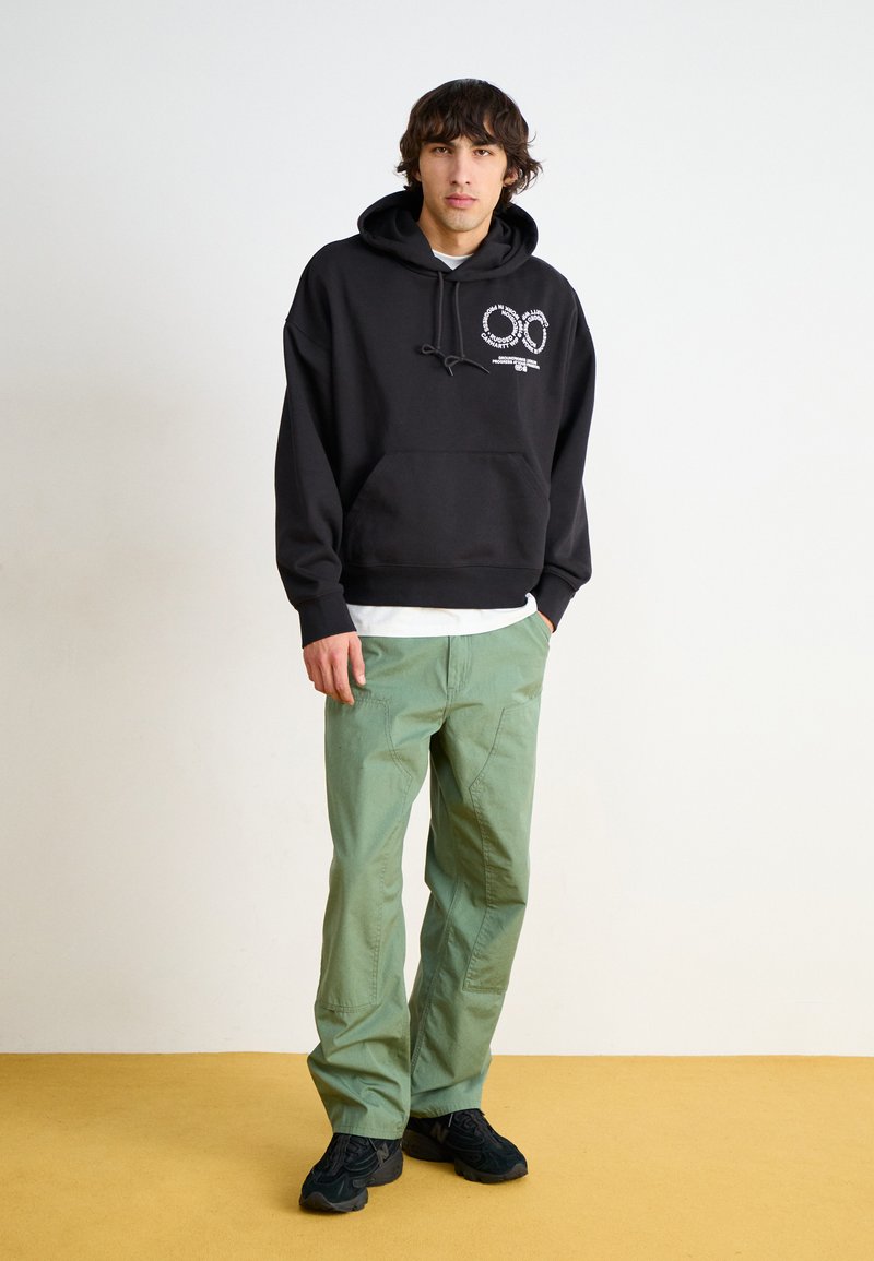 Sudadera negra con un gráfico frontal, de material de algodón, combinada con pantalones cargo verdes que tienen múltiples bolsillos y un ajuste relajado, usados con zapatillas oscuras.