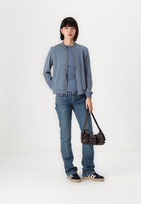 Lys blå bomberjakke, tettsittende blå t-skjorte, denimjeans, sorte joggesko med hvite striper, som holder en liten mørkebrun håndveske.