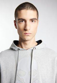 Šedá mikina s kapucí s výrazným šedým motivem lebky na přední straně, vybavena šňůrkovou kapucí a měkkou, texturovanou tkaninou.