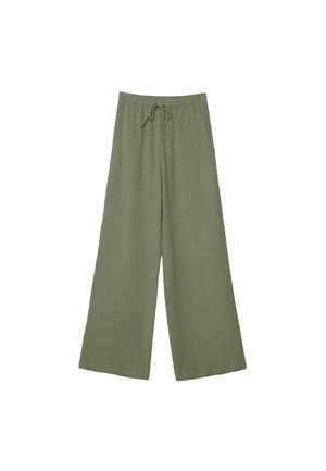 Mladá žena stojaca v širokých olivovo zelených nohaviciach a krémovom bezrukávovom na záhyboch topánke na jednoduchom svetlom pozadí.