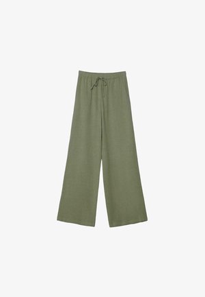 Mladá žena stojaca v širokých olivovo zelených nohaviciach a krémovom bezrukávovom na záhyboch topánke na jednoduchom svetlom pozadí.