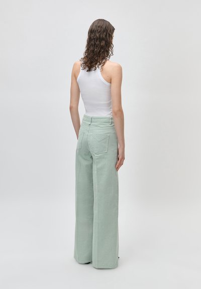 Γυναίκα με καστανά κατσαρά μαλλιά, ντυμένη με λευκό αμάνικο τοπ και υψηλόμεσα, φαρδιά παντελόνια μέντας, που στέκεται γυρισμένη με την πλάτη.