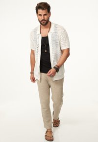 Weißes offenes Strickshirt mit kurzen Ärmeln über einem schwarzen Top, kombiniert mit beigen strukturierten Hosen und braunen Sandalen, accessoriert mit Armbändern.