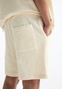Shorts couleur crème en tissu texturé. Présentent une poche latérale avec une petite étiquette de marque. L'ourlet est droit et sans décoration.