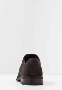 Chaussure en daim marron avec une texture lisse, une forme arrondie et une semelle sombre contrastante avec des rayures subtiles. Vue arrière affichée.