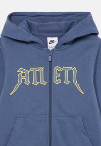 Felpa con zip Nike di colore blu scuro, con un testo grafico giallo "ATLET" sul petto. Include un cappuccio e tasche frontali.