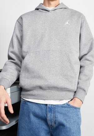 Person trägt grauen Kapuzenpullover mit weißem Logo auf der Brust, blaue Jeans, steht neben einem Drucker.