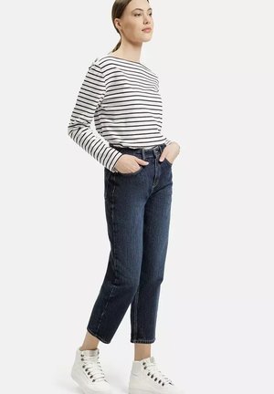 Kobieta ubrana w białą i czarną striped koszulę z długim rękawem, ciemne spodnie typu cropped, białe sneakersy za kostkę, stojąca z rękami w kieszeniach.