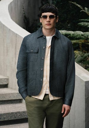 Nuori mies aurinkolaseissa, tummanharmaa napitettu takki, beige ruudullinen paita ja oliivinvihreät housut, seisomassa betonisten portaiden ja kasvillisuuden vieressä.