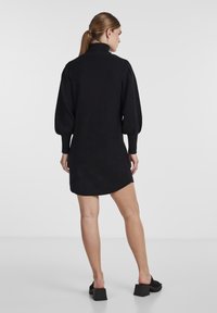Robe-sweat noire avec col haut, longues manches bouffantes et ourlet incurvé. Fabriquée en tissu doux, portée avec des chaussures à talons blocs noires.