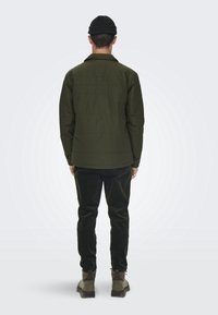 Veste rembourrée verte avec un col, en tissu texturé et coupe droite, associée à un pantalon en velours côtelé vert foncé et des bottes marron.
