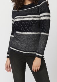 Svart och grå stickad tröja med horisontella och diamantmönster, rund halsringning och långa ärmar med vita ränder vid muddar och fåll.