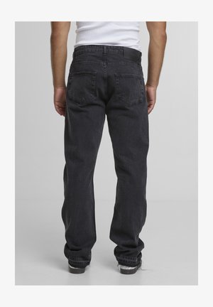 Person trägt schwarze Jeans mit geradem Bein und Gesäßtaschen sowie ein weißes ärmelloses Top, steht vor einem einfarbigen grauen Hintergrund.