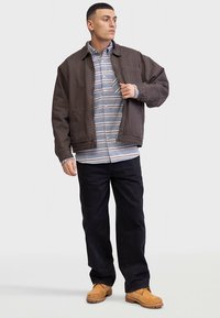 Pruun bomberjakk sinise ja valge triibulise särgi peal, kombineeritud mustade pükste ja beežide kingadega. Riietus on lõdvestunud lõikega ja tekstuurse kangaga.