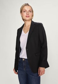 Schwarze Blazer mit einem einzigen Knopf, taillierte Passform und feinen Revers, getragen über einem weißen V-Ausschnitt-Shirt und dunkelblauen Jeans. Glatte Textur.