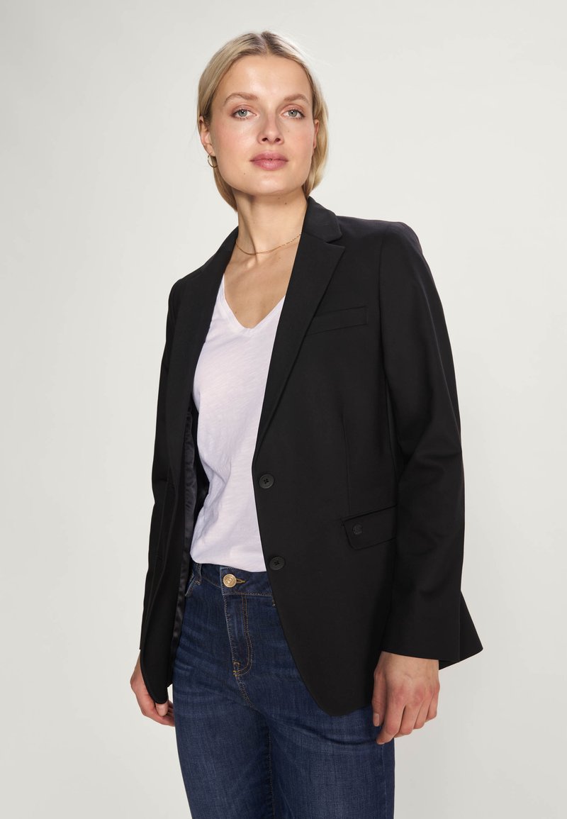 Schwarze Blazer mit einem einzigen Knopf, taillierte Passform und feinen Revers, getragen über einem weißen V-Ausschnitt-Shirt und dunkelblauen Jeans. Glatte Textur.