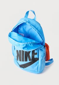 Ανοιχτό μπλε σακίδιο πλάτης Nike με μεγάλο μαύρο λογότυπο, ανοιχτό κύριο διαμέρισμα και μικρή πορτοκαλί πλαϊνή θήκη σε λευκό φόντο.